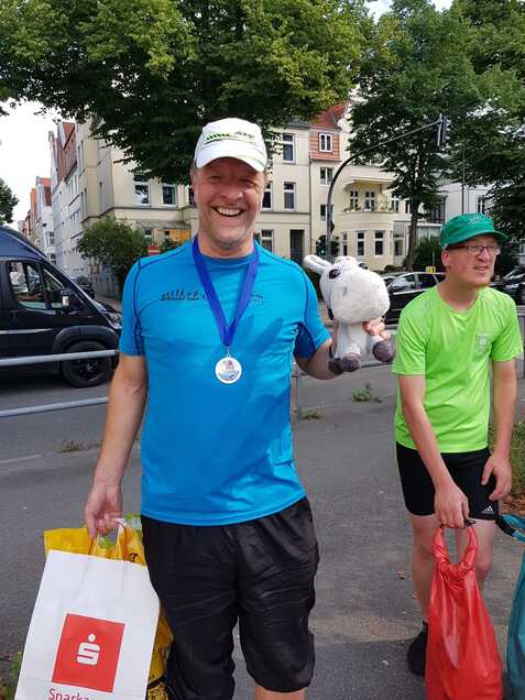 Lachender Mann in blauer Sportkleidung hält ein Stofftier und eine Tasche, im Hintergrund sind Bäume und Gebäude.