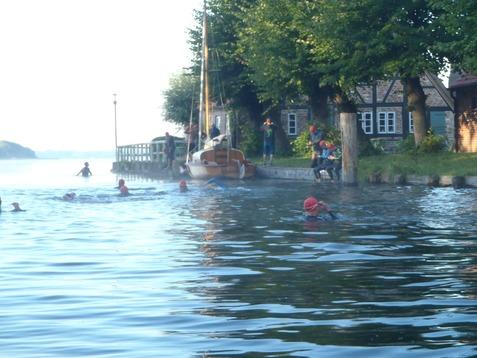 Schwimmer in roten Badekappen im Wasser neben einem Bootssteg mit einem Segelboot und Dächer im Hintergrund.
