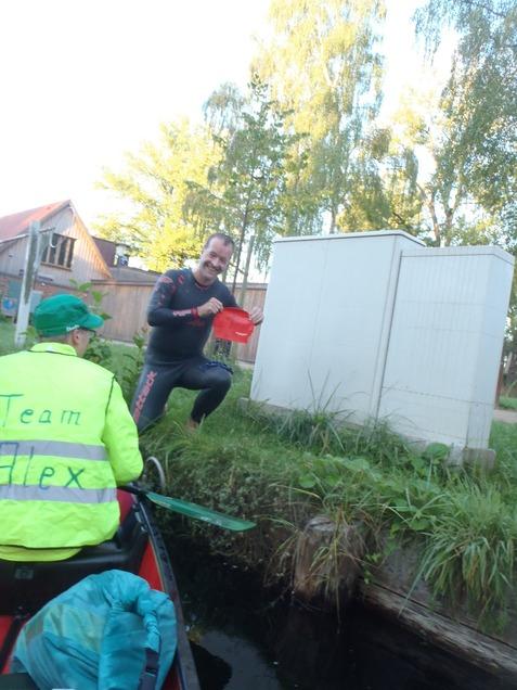 Ein Mann in einem Neoprenanzug hält eine rote Schwimmkappe, während ein anderer im Boot sitzt. Hintergrund mit Bäumen.