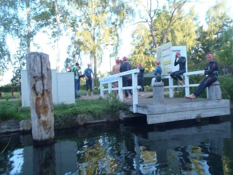Gruppierung von Schwimmern in Neoprenanzügen und Badekappen auf einem Steg am Wasser, umgeben von Bäumen und Wiesen.