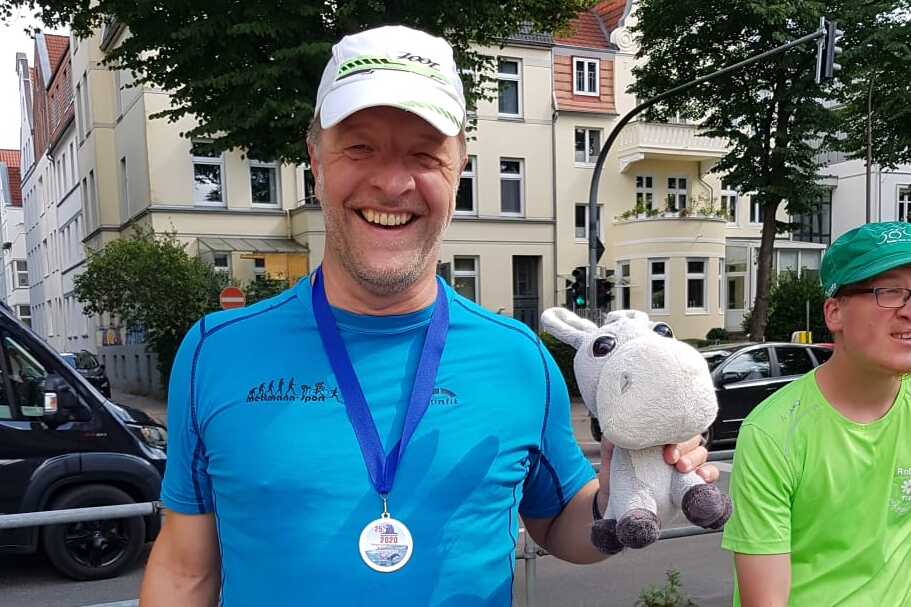 Lachender Mann in blauer Sportkleidung hält ein Stofftier und eine Tasche, im Hintergrund sind Bäume und Gebäude.
