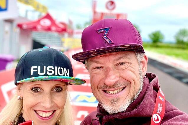 Paar mit Medaillen posiert lächelnd an einer Rennstrecke, im Hintergrund Werbebanner und grüne Landschaft.