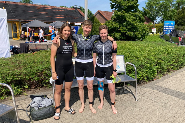 Drei Athletinnen in Sportanzügen stehen lächelnd auf einer Bank, umgeben von grünen Sträuchern und Wettkampfatmosphäre.