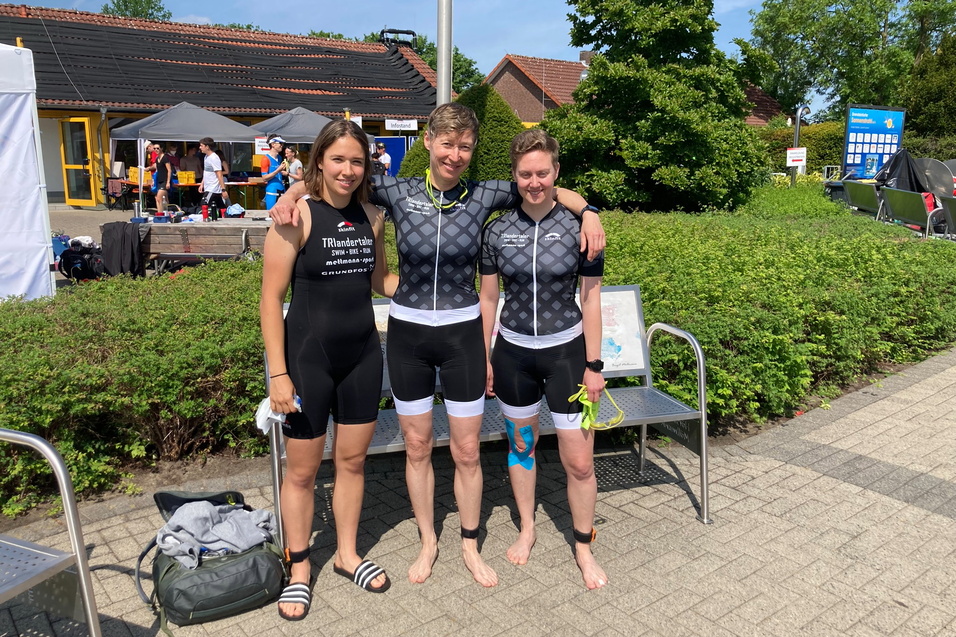 Drei Athletinnen in Sportanzügen stehen lächelnd auf einer Bank, umgeben von grünen Sträuchern und Wettkampfatmosphäre.