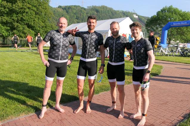 Vier sportlich gekleidete Männer in Wettkampfbekleidung stehen lachend auf einer Rasenfläche vor einer Veranstaltung.