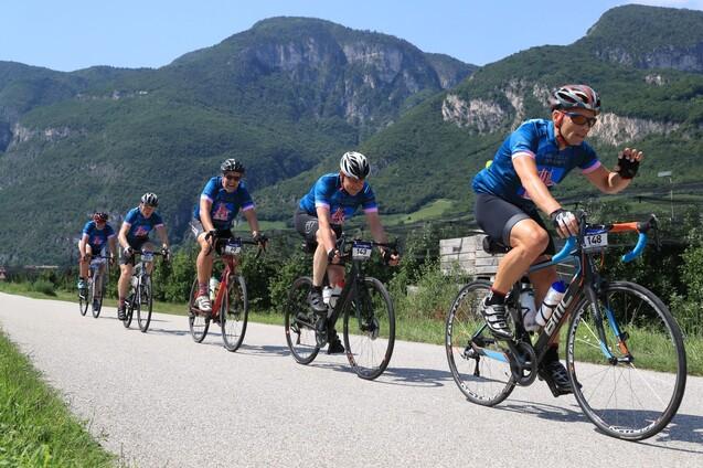 Sechs Radfahrer in blauen Trikots fahren auf einer Straße mit Bergen im Hintergrund.