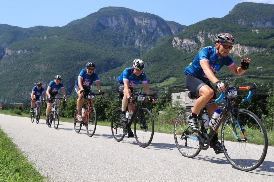 Sechs Radfahrer in blauen Trikots fahren auf einer Straße mit Bergen im Hintergrund.