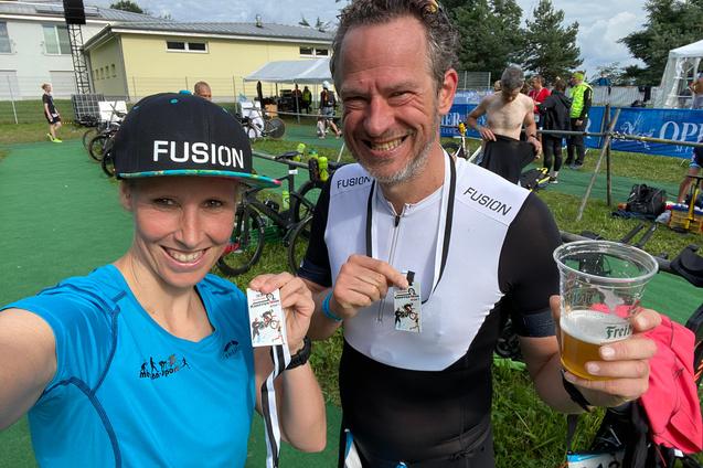 Selbstporträt zweier Personen mit Medaillen in der Hand, im Hintergrund Fahrräder und Teilnehmer eines Sportevents.