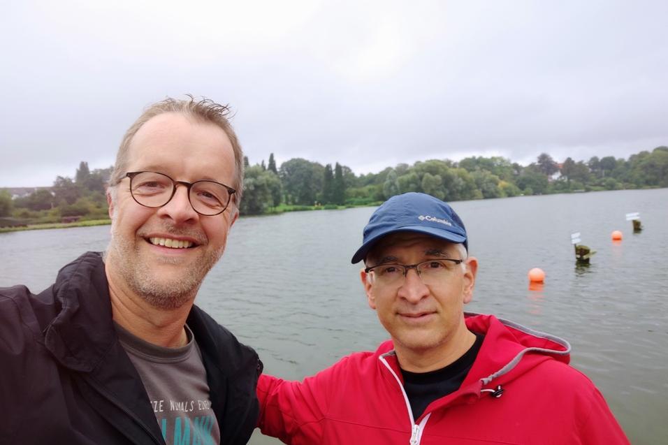 Zwei Männer posieren lächelnd am Ufer eines Sees, umgeben von grünem Uferbewuchs und einer grauen Himmelstimmung.