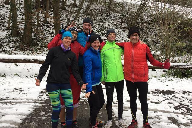 Sechs Personen posieren im Schnee; einige tragen Laufbekleidung und zeigen fröhliche Gesten.