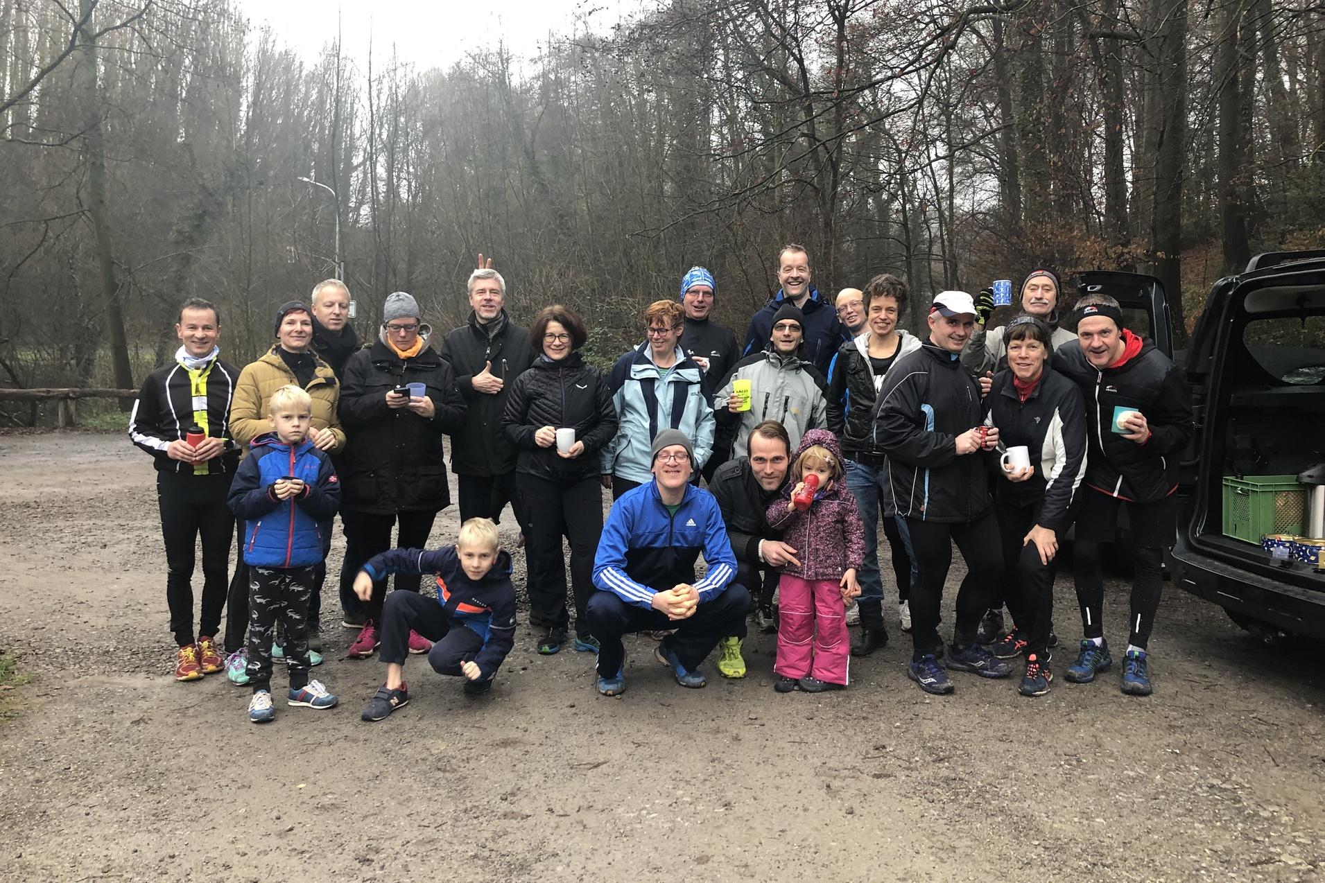 Gruppe von Läufern in Sportkleidung posiert in einem Waldgebiet, einige halten Tassen in den Händen.