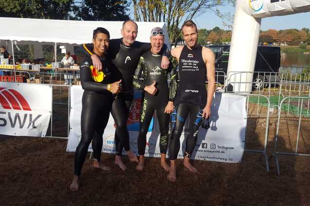 Vier Schwimmer in Neoprenanzügen stehen lächelnd vor einem Zelt am Wasser, bereit für einen Wettkampf.