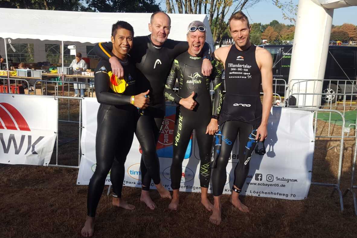 Vier Schwimmer in Neoprenanz&uuml;gen stehen l&auml;chelnd vor einem Zelt am Wasser, bereit f&uuml;r einen Wettkampf.