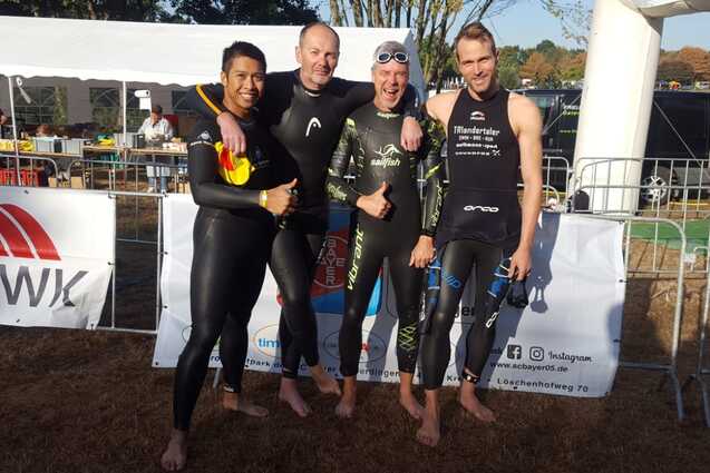 Vier Schwimmer in Neoprenanz&uuml;gen stehen l&auml;chelnd vor einem Zelt am Wasser, bereit f&uuml;r einen Wettkampf.