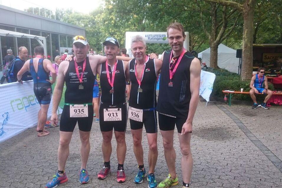 Vier Männer in Sportkleidung stehen Arm in Arm, tragen Medaillen und Finisher-Medaillen, im Hintergrund Teilnehmer und Stände.