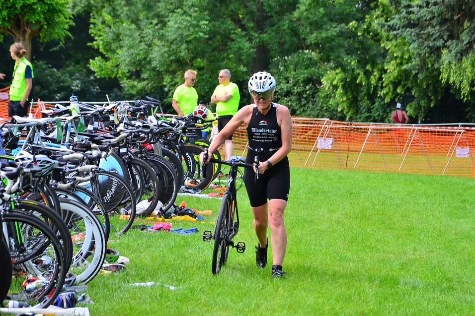 Eine Triathletin in schwarz-weißer Sportbekleidung schiebt ihr Fahrrad über grüne Wiese, umgeben von anderen Fahrrädern.