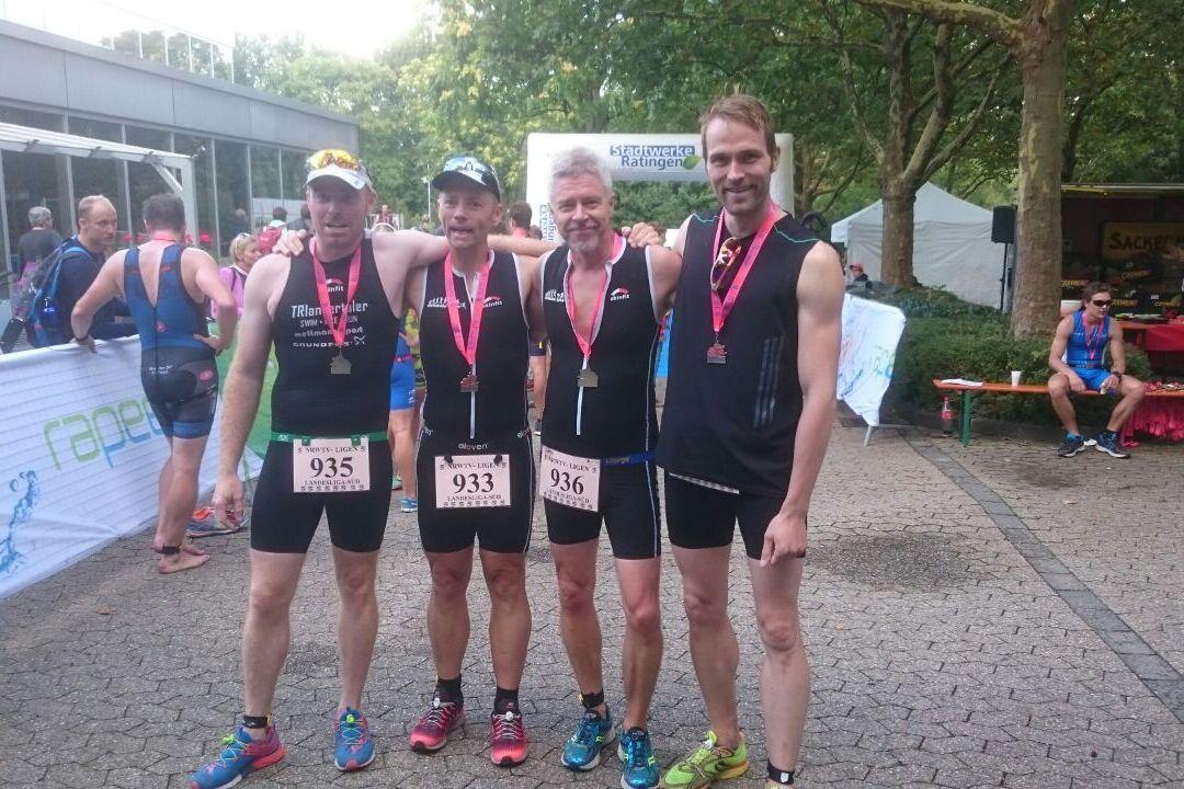 Vier Männer in Sportkleidung stehen Arm in Arm, tragen Medaillen und Finisher-Medaillen, im Hintergrund Teilnehmer und Stände.