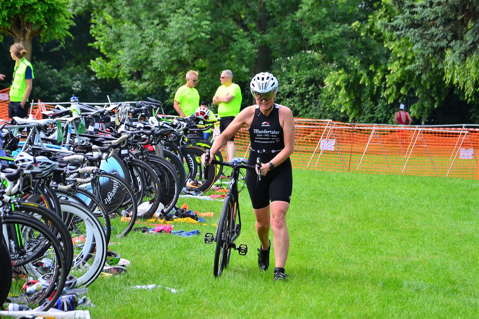 Eine Triathletin in schwarz-weißer Sportbekleidung schiebt ihr Fahrrad über grüne Wiese, umgeben von anderen Fahrrädern.