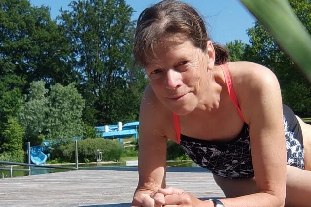 Frau in Badebekleidung macht eine Planke auf einem Holzdeck, im Hintergrund ist eine grüne Landschaft und Wasserrutschen zu sehen.