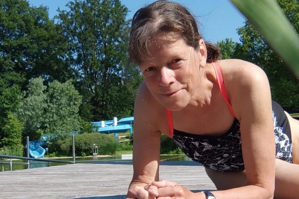 Frau in Badebekleidung macht eine Planke auf einem Holzdeck, im Hintergrund ist eine grüne Landschaft und Wasserrutschen zu sehen.