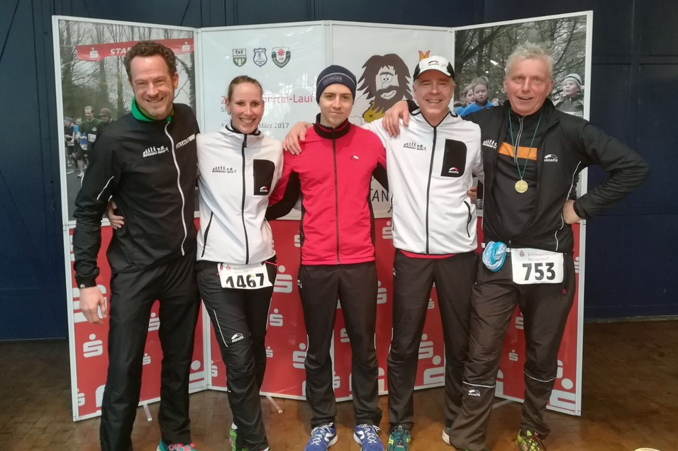 Fünf Läufer posieren lächelnd vor einem Banner bei einer Laufveranstaltung, ihre Startnummern sind sichtbar.