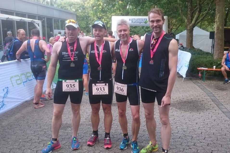 Vier Sportler in Triathlonausrüstung stehen fröhlich mit Medaillen um den Hals nach dem Wettkampf zusammen.