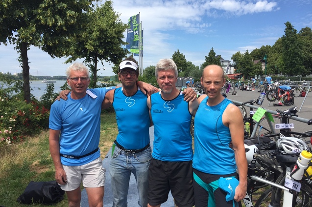 Vier Männer in blauen Sporttrikots stehen nebeneinander an einem Gewässer, mit Fahrrädern im Hintergrund.