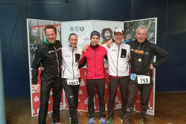 Fünf Läufer posieren lächelnd vor einem Banner bei einer Laufveranstaltung, ihre Startnummern sind sichtbar.