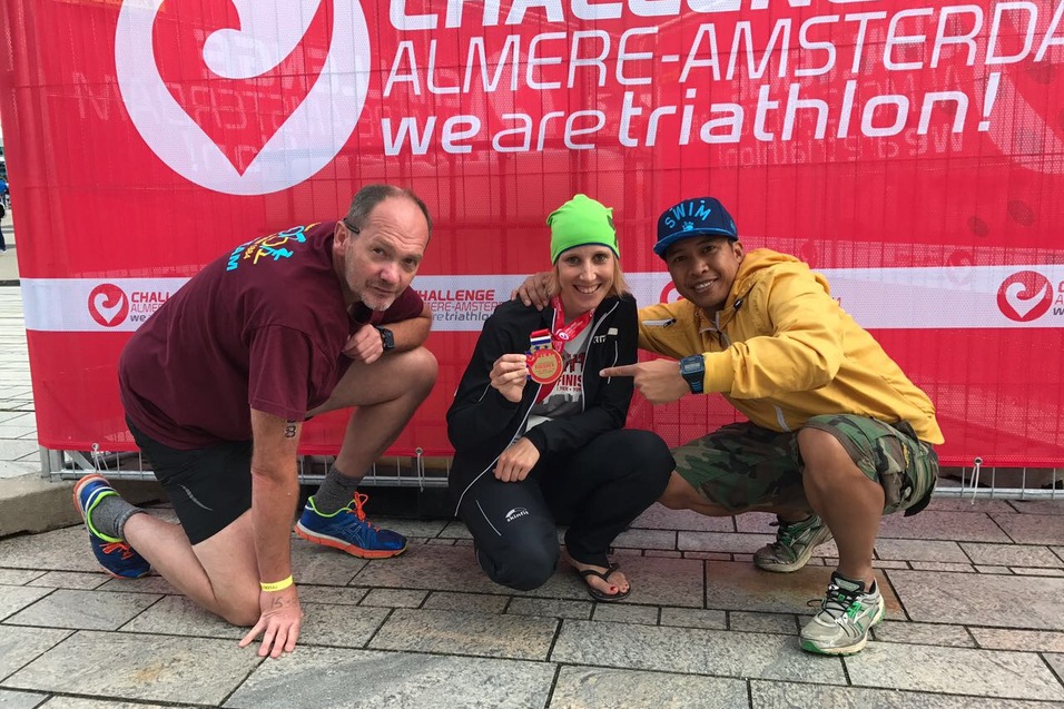 Drei Personen posieren lächelnd vor einem roten Banner nach einem Triathlon, eine hält eine Medaille in der Hand.