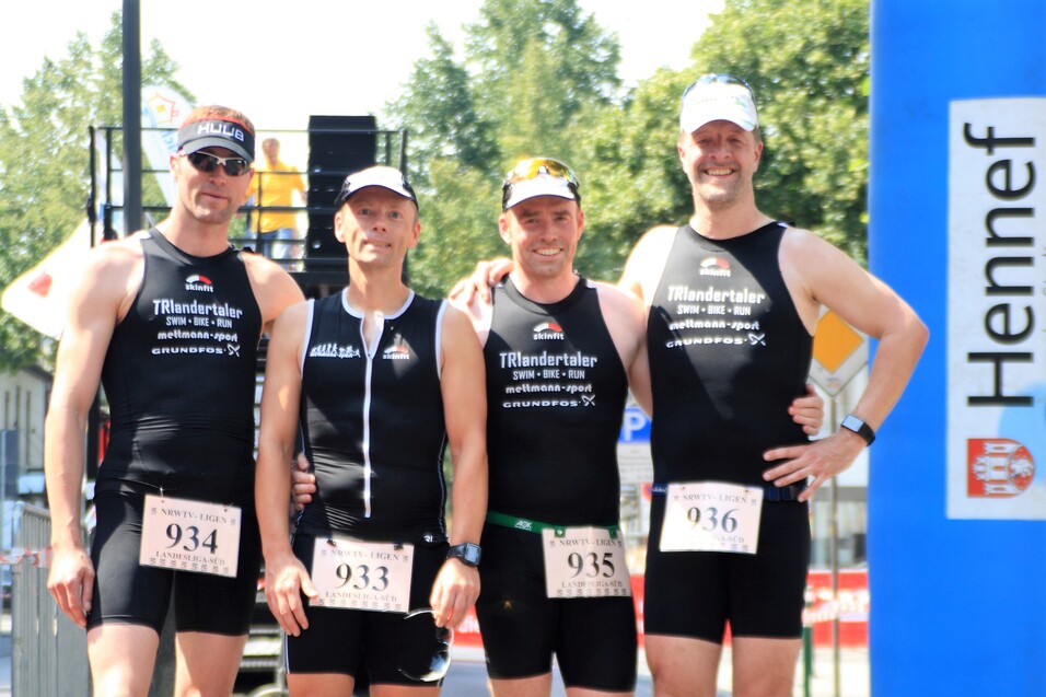 Vier Männer in Sportanzügen stehen lächelnd vor einem Event-Hintergrund, bereit für einen Triathlon-Wettkampf.