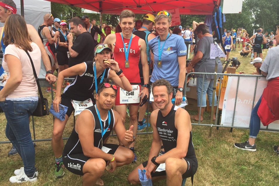Gruppe von Triathleten in Sportkleidung posiert lächelnd vor einem Wettkampfbereich mit Zuschauern im Hintergrund.