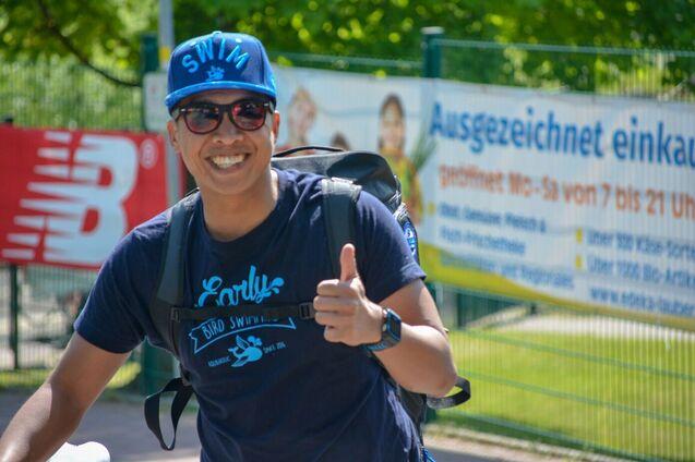 Lächelnder Mann mit Sonnenbrille und Baseballmütze, zeigend Daumen hoch, im Freien vor einem Werbebanner.