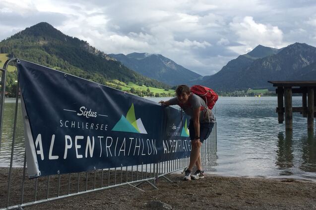 Person mit Rucksack beugt sich über eine Absperrung am Schliersee, im Hintergrund Berge und ein Steg sichtbar.