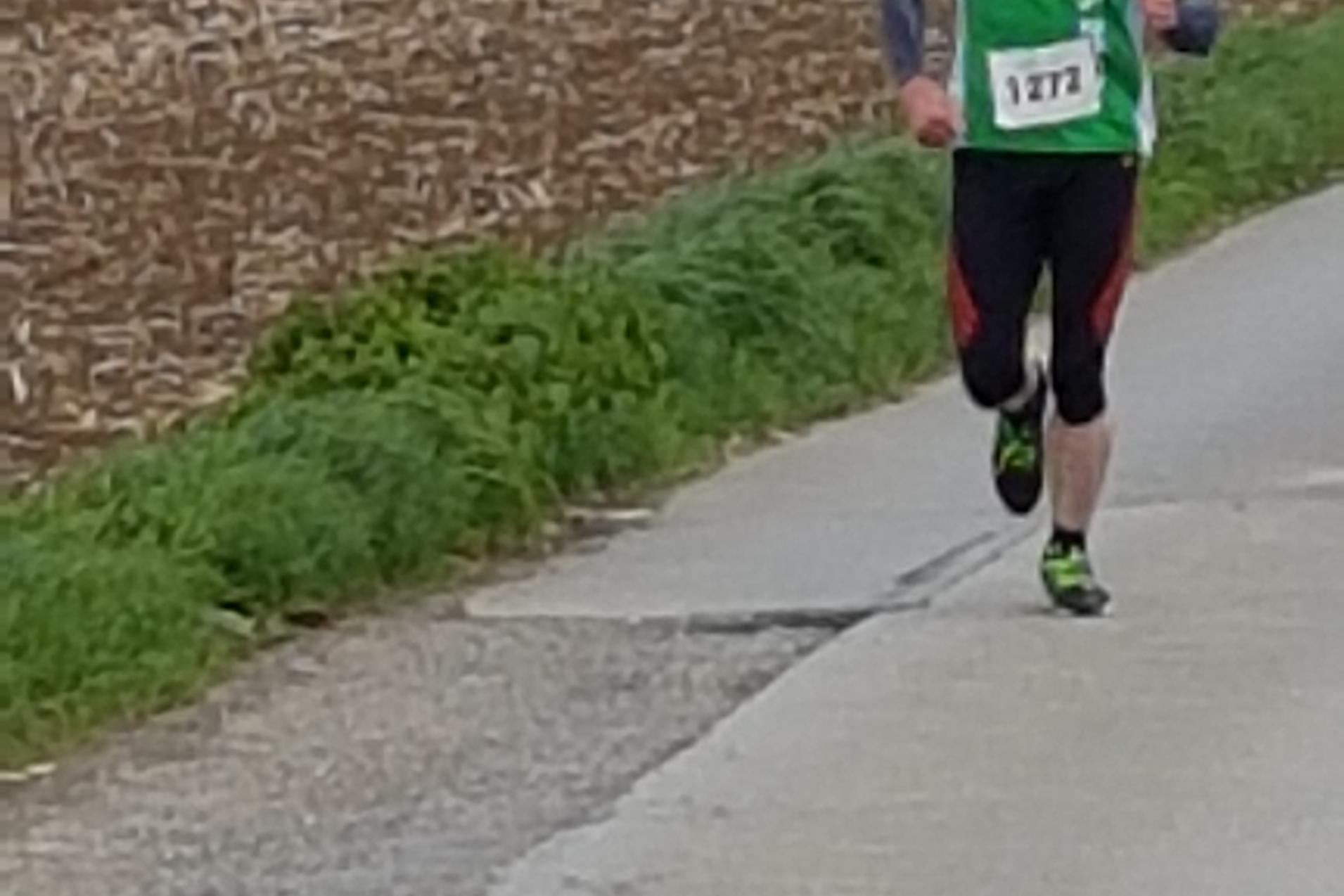 Läufer in grünem Shirt mit Startnummer 372 joggt auf einer Straße, umgeben von einem Feld und Bäumen im Hintergrund.