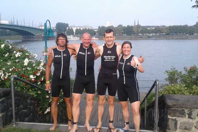 Vier sportlich gekleidete Personen in Neoprenanzügen lächeln am Ufer eines Flusses mit Brücke im Hintergrund.