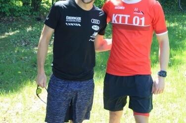Zwei Männer in Sportkleidung stehen lächelnd im Freien vor grüner Natur. Einer trägt ein schwarzes, der andere ein rotes Shirt.