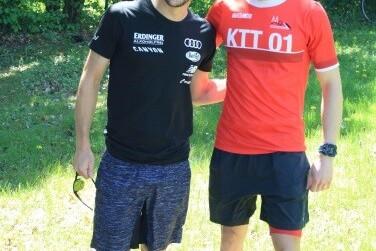 Zwei Männer in Sportkleidung stehen lächelnd im Freien vor grüner Natur. Einer trägt ein schwarzes, der andere ein rotes Shirt.