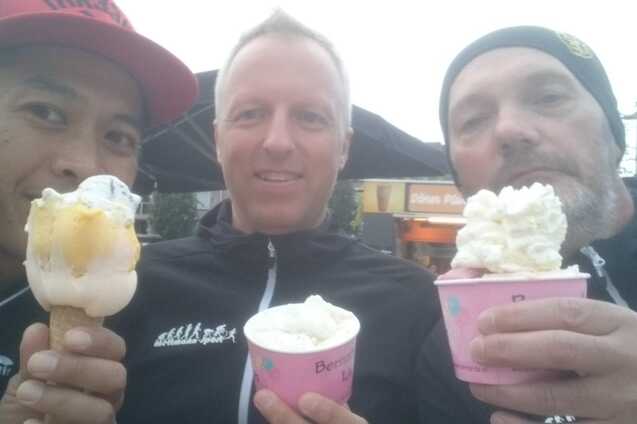 Drei Männer halten Eisbecher und Hörnchen, lächeln in die Kamera vor einem Outdoor-Café.