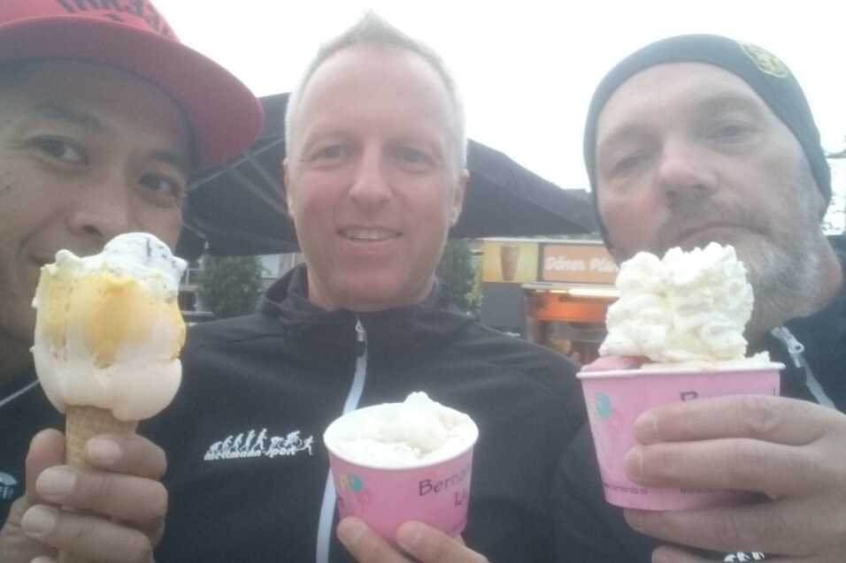 Drei Männer halten Eisbecher und Hörnchen, lächeln in die Kamera vor einem Outdoor-Café.