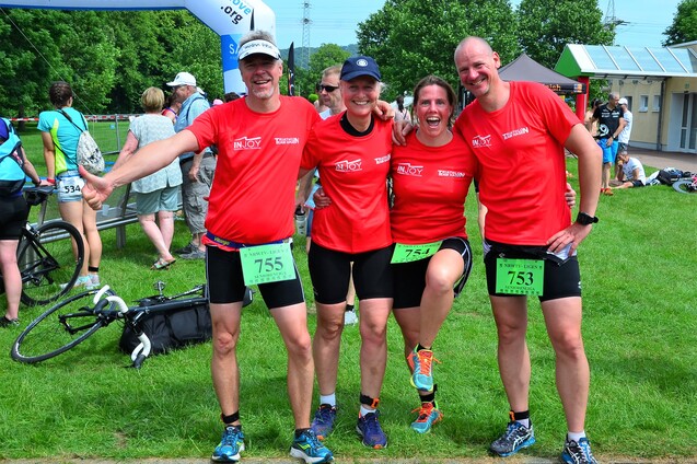 Vier Personen in roten Shirts stehen lächelnd auf einer Wiese, mit Startnummern an den Beinen und Fahrrädern im Hintergrund.