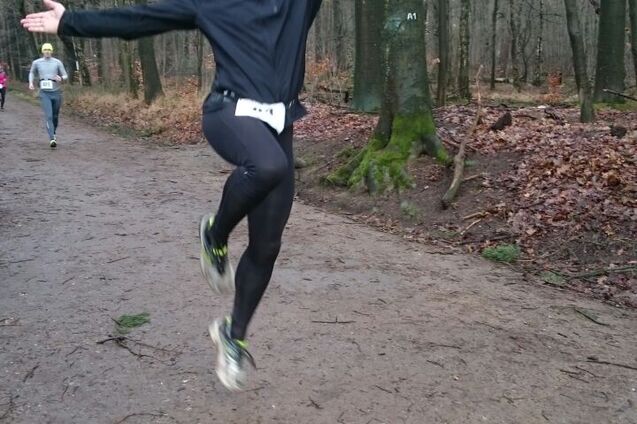 Läufer springt fröhlich auf einem Waldweg, umgeben von Bäumen, mit anderen Athleten im Hintergrund.