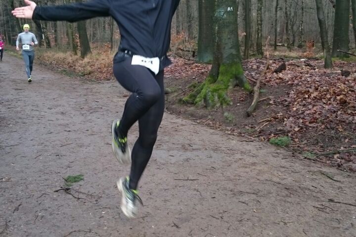 Läufer springt fröhlich auf einem Waldweg, umgeben von Bäumen, mit anderen Athleten im Hintergrund.
