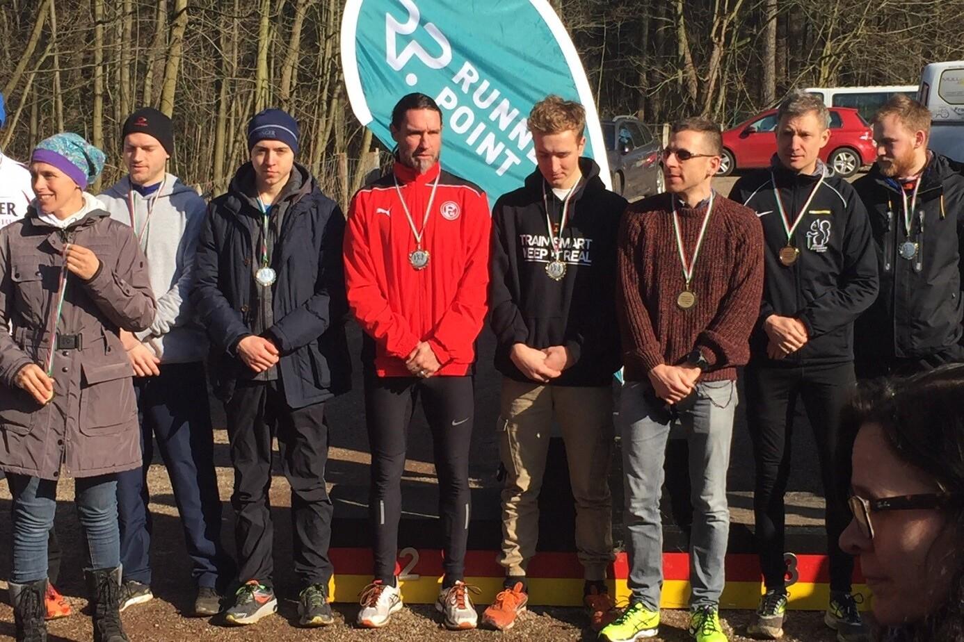Gruppe von Läufern mit Medaillen bei einer Siegerehrung vor einem blauen Banner im Wald.
