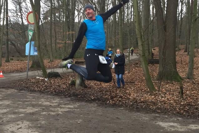 Ein Läufer springt begeistert in einem Wald, während Zuschauer im Hintergrund die Strecke beobachten.