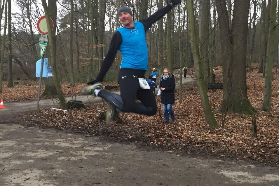 Ein Läufer springt begeistert in einem Wald, während Zuschauer im Hintergrund die Strecke beobachten.