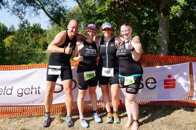 Vier Triathleten in Wettkampfanzügen posieren lächelnd mit Daumen hoch vor einer Banner-Wand und grünem Hintergrund.