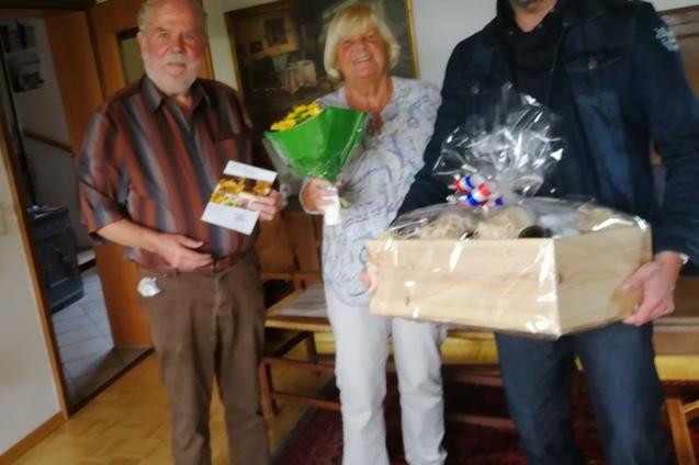 Drei Personen stehen in einem Raum, eine hält einen Blumenstrauß, die anderen Geschenke und eine Broschüre.