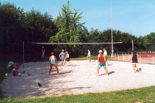 Gruppe von Personen spielt Beachvolleyball auf einem Sandplatz im Freien, umgeben von Bäumen und Sportanlagen.