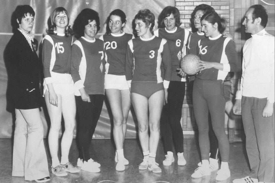 Gruppe weiblicher Sportlerinnen in Trikots mit Nummern, posierend in einer Halle mit einem Basketball.