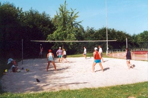 Gruppe von Personen spielt Beachvolleyball auf einem Sandplatz im Freien, umgeben von Bäumen und Sportanlagen.